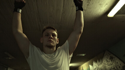 A young athlete in a white t-shirt and boxing gloves stands victoriously in a dimly lit gym, arms raised, embodying resilience and warrior spirit in a gritty combat sports setting.