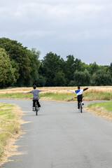 Fototapeta premium Two boys riding bicycles with arms outstretched on a rural road surrounded by fields and trees. 