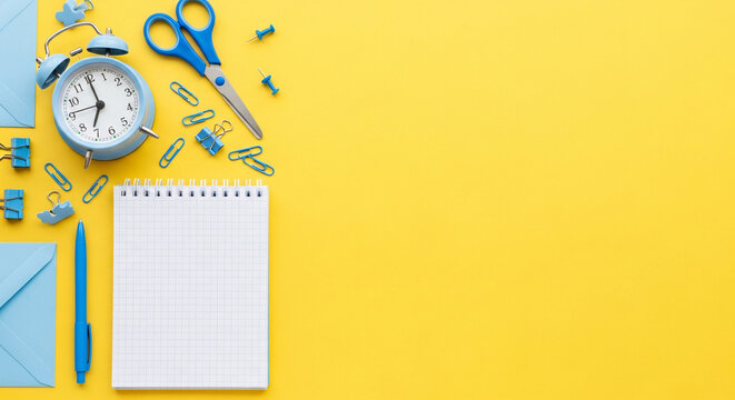 Organized workspace essentials including a retro alarm clock, scissors, notebook, pen, paper clips, and envelopes laid out on a vibrant yellow background for productivity planning. - Powered by Adobe