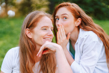 Two women mother and daughtershare a secret while laughing during a sunny day in a lush green park,...