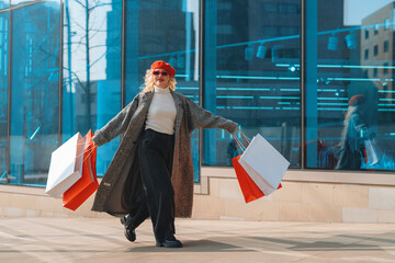 Woman wearing red beret and sunglasses joyfully carries shopping bags while walking outside modern...
