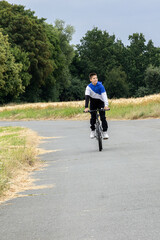 Fototapeta premium Boy riding a bicycle on a rural road with green trees and fields around. 