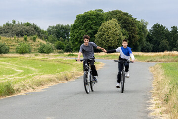 Obraz premium Two boys riding bicycles holding hands on a rural road surrounded by fields and trees. 