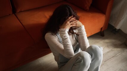 Depressed young woman sitting on floor near sofa, hands covering face, experiencing intense emotional distress and overwhelming mental anguish in quiet home environment - Powered by Adobe