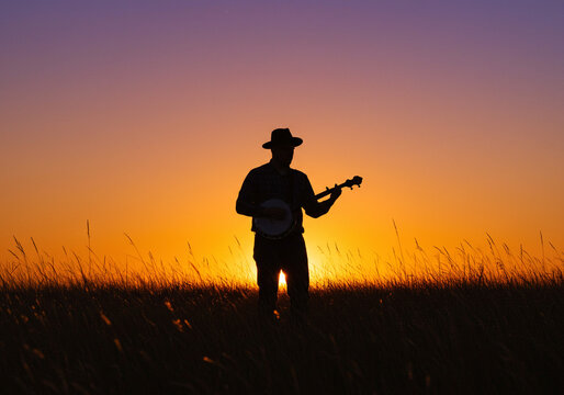 Silhouette of a musician playing banjo at sunset in a field with orange and purple sky backdrop - Powered by Adobe