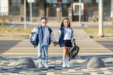 Happy little boy and girl, friends, both schoolchildren, stand hand in hand in front of modern school building, hold backpacks. Children friendship, companionship and the beginning of a school day.