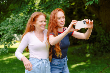 Friends mother, daughter enjoying a sunny day outdoors while taking selfies in a lush green park...