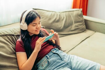 Asian teen girl on green sofa using smartphone with headphones, focused expression home interior, light from window technology gadgets, lifestyle