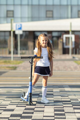 Happy little girl holding backpack, stands near scooter in front of modern school building. Child...