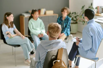 Fotobehang Group of multiethnic teenagers sitting in circle participating in discussion with Caucasian male counselor holding clipboard during therapy or support session indoors © AnnaStills