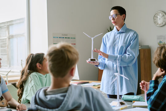 Middle aged Caucasian woman explaining wind turbine model to group of teenagers in classroom setting, students listening attentively while observing renewable energy demonstration