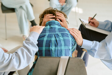 Teenager sitting with head in hands surrounded by supportive group, hands resting on shoulders, counselor taking notes during therapy session