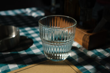 Clear glass of water on checkered tablecloth with sunlight and shadows, evoking freshness and calm atmosphere in still life composition.