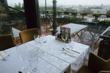 Empty restaurant table set with glassware and cutlery on rainy day overlooking outdoor terrace with metal chairs and city view beyond.
