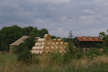 Rural landscape with stacked hay bales, old wooden barns, green trees, and cloudy sky in a peaceful countryside setting.