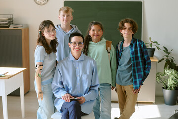 Group of diverse teenagers standing with Caucasian middle aged woman wearing glasses in classroom,...