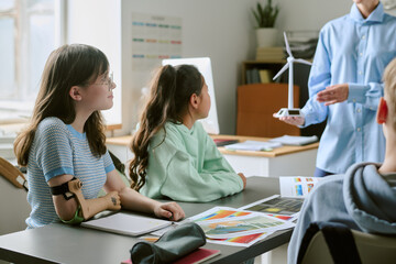 Caucasian teenage girl with prosthetic arm sitting at desk listening to teacher presenting wind turbine model, multiethnic teenagers engaging in classroom science lesson