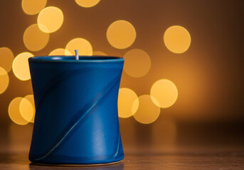 A blue candle in a ceramic holder with a blurred background of yellow lights on a wooden surface