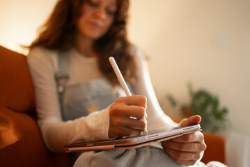 Concentrating on a digital art project, a female artist sits comfortably on a sofa, skillfully using a stylus and tablet to bring her creative vision to life