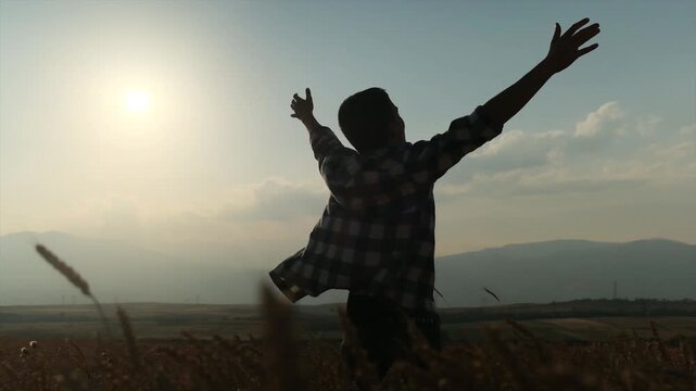Silhouette of a young man in an open shirt standing in a wheat field at sunset, raising his arms in joy among high mountains. A happy, life-loving person enjoying a moment of freedom and peace.