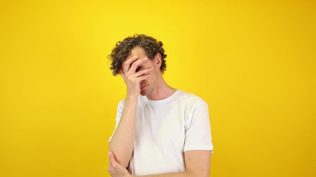 Shocked man covers his face with hands, peering fearfully through fingers against a yellow studio backdrop - perfect for Halloween promotions, horror films, and mental health awareness.