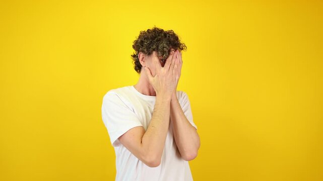 Shocked man covers his face with hands, peering fearfully through fingers against a yellow studio backdrop - perfect for Halloween promotions, horror films, and mental health awareness.