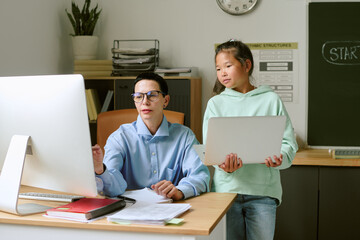 Middle aged Caucasian woman sitting at desk using desktop computer while Asian girl teenager...