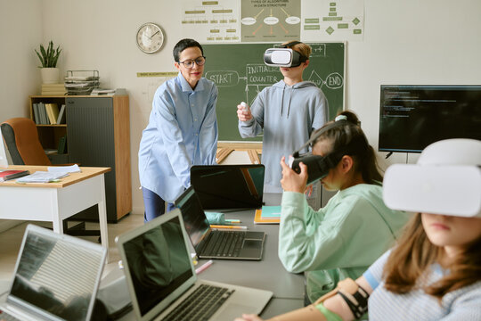 Asian middle aged woman teaching group of teenagers using virtual reality headsets and laptops in classroom, students interacting with technology during educational activity