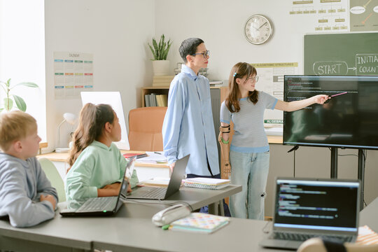 Teenage boy and teenage girl standing near interactive display, girl pointing at screen while two Caucasian children sitting at desks with laptops watching presentation