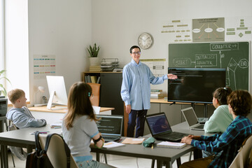 Caucasian middle aged woman teaching group of teenagers coding in classroom, standing near digital screen with programming code, students sitting at desks using laptops and listening