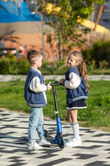 Sibling children with school backpack stand with scooter on the background of modern city park....