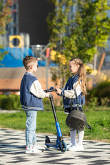 Sibling children with school backpack stand with scooter on the background of modern city park....