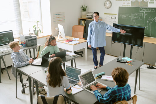Caucasian middle aged woman teaching group of teenagers in classroom, students using laptops while listening and taking notes, teacher pointing at large monitor near chalkboard