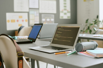 Empty classroom with two open laptops on desks displaying programming code, notebooks and stationery scattered around, modern educational environment suggesting technology learning setting