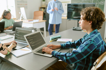 Teenage Caucasian boy working on laptop with code on screen in classroom, surrounded by classmates including Asian girl and student with prosthetic arm, teacher standing near chalkboard