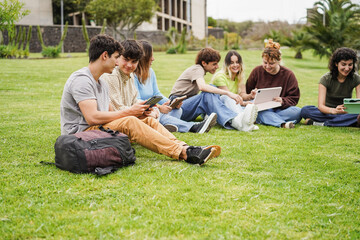 Happy students studying with digital tablets sitting on grass at campus park with college building in background - Back to school concept - Main focus on left boy face