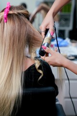 Fototapeta premium Hair stylist curling blonde hair with a flat iron in a beauty salon. Back view of a woman with half-straight and half-curled hair during hair styling process. Hands with hair tool visible