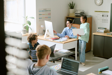 Group of teenagers working together in modern classroom, teacher explaining project on computer to Asian girl while two other teenagers using laptops in foreground