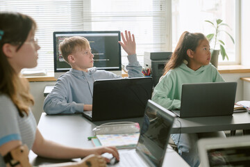 Group of teenagers including Caucasian boy raising hand and Asian girl sitting at desks using laptops in classroom setting, engaging in learning activity with focused expressions