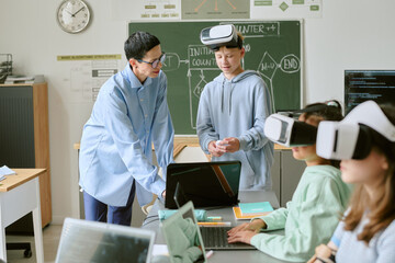 Female teacher instructing group of teenagers using virtual reality headsets in classroom, students engaging with technology while teacher explaining concept near chalkboard