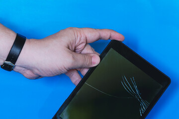 Hand holding tablet with cracked screen on blue plain background, broken tablet screen held by hand, damaged tablet in hand.