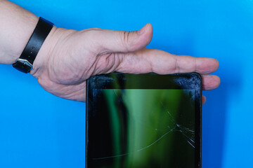 Hand pressing old tablet with cracked screen on blue plain background, broken tablet screen held by hand, disposal of broken old devices.