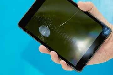 Hand holding broken and cracked tablet with fingerprint on the crack, hand holding old tablet with cracked screen on blue plain background close-up, disposal of broken old devices.