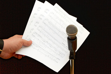 Hand holding sheet music next to microphone on microphone stand on black background, close-up, music notes for performer in front of microphone, backdrop for podcast, cover for audio podcast.