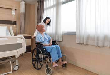 A caregiver support a chemo patient with knitted hat on her head sitting in wheelchair, they enjoy the natural light coming through the window in hospital room. The atmosphere is calm and comforting.