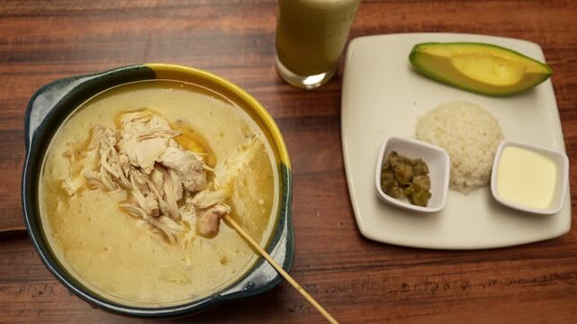 Overhead view of creamy ajiaco soup paired with rice, avocado, and flavorful sides on a wooden table. Warm lighting enhances this enticing Colombian dish presentation.