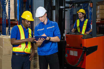 Two warehouse worker in hard hat and safety vest discuss operational detail while examining document in an industrial setting. They focus intently on their task.