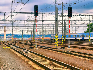 Fototapeta premium Railway Tracks and Overhead Power Lines at Train Station – Abstract Industrial Background with Red Signals and Blue Passenger Train, Ideal for Wallpaper, Web Design, Advertising, and Digital Use