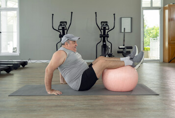Senior chubby guy doing physical exercise with fitness ball lying on floor at gym. Side view of man having sport workout after gaining weight and getting fat.