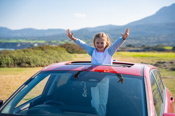 Little girl joyfully sticks out of car sunroof with raised hands against the backdrop of mountains in Northern Cyprus. The image symbolizes freedom, adventure, and the excitement of travel with child